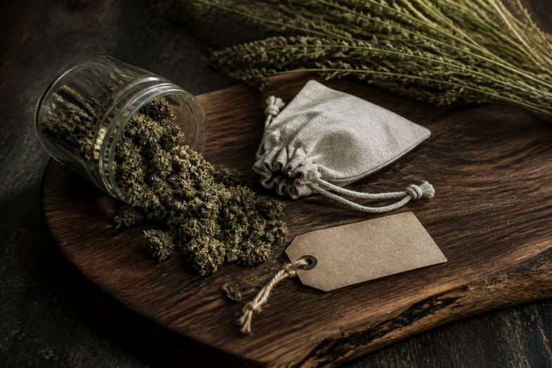 Getrocknete CBD-Blüten fallen aus einem umgekippten Glas auf ein dunkles Holzbrett, daneben ein Leinensäckchen